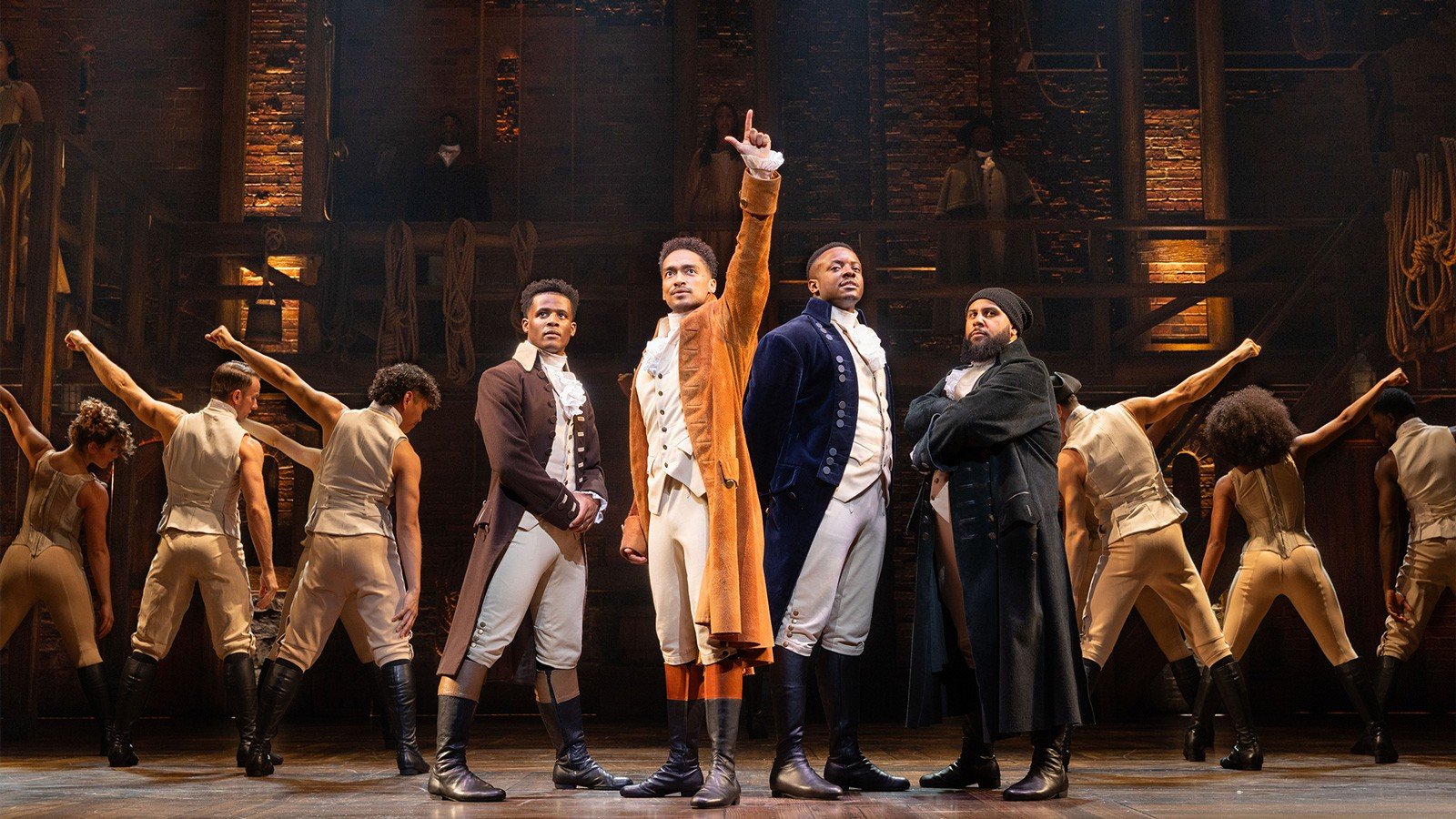 Production image from Hamilton the musical in London. Hamilton stands one arm upstretched, the cast fanning out behind him in a triangle.